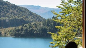 A person sitting by a window reading a book, with a view of a lake and forested hills. - Hermoso Eco Lodge (San Martín de los Andes)