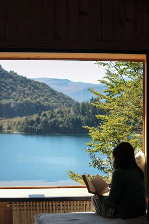 A person sitting by a window reading a book, with a view of a lake and forested hills. - Hermoso Eco Lodge (San Martín de los Andes)