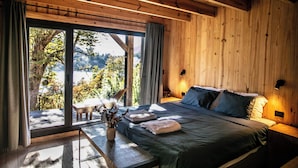 A wooden room with a bed, a table with a vase, and a view of trees and mountains through large windows. - Hermoso Eco Lodge (San Martín de los Andes)