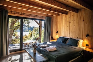 A wooden room with a bed, a table with a vase, and a view of trees and mountains through large windows. - Hermoso Eco Lodge (San Martín de los Andes)