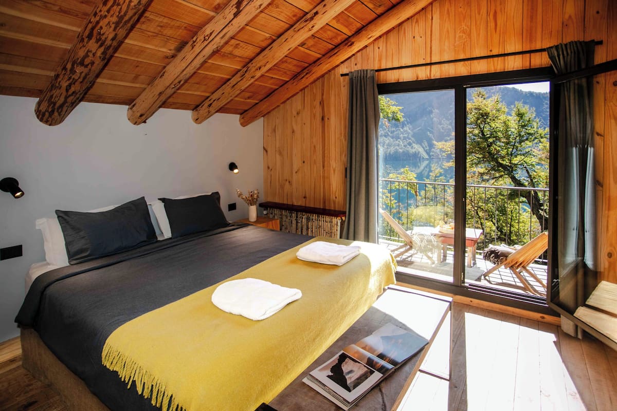 A bedroom with a large bed, a wooden ceiling, a balcony with a view of trees and mountains, and a window with curtains.