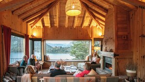 A wooden cabin interior with a fireplace, large windows overlooking a lake, and a group of people seated on a couch. - Hermoso Eco Lodge (San Martín de los Andes)