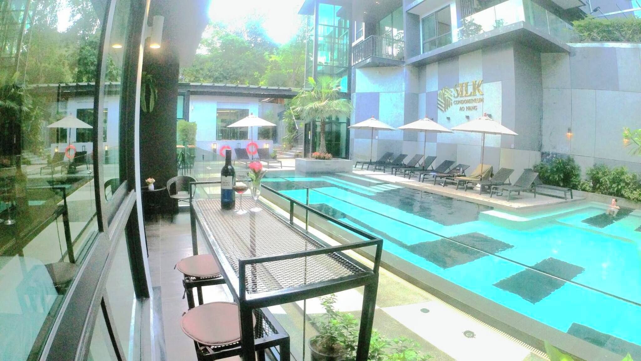 A swimming pool with lounge chairs and umbrellas, a table with chairs, and a building with a sign reading "SILK CONDOMINIUM AO NANG".