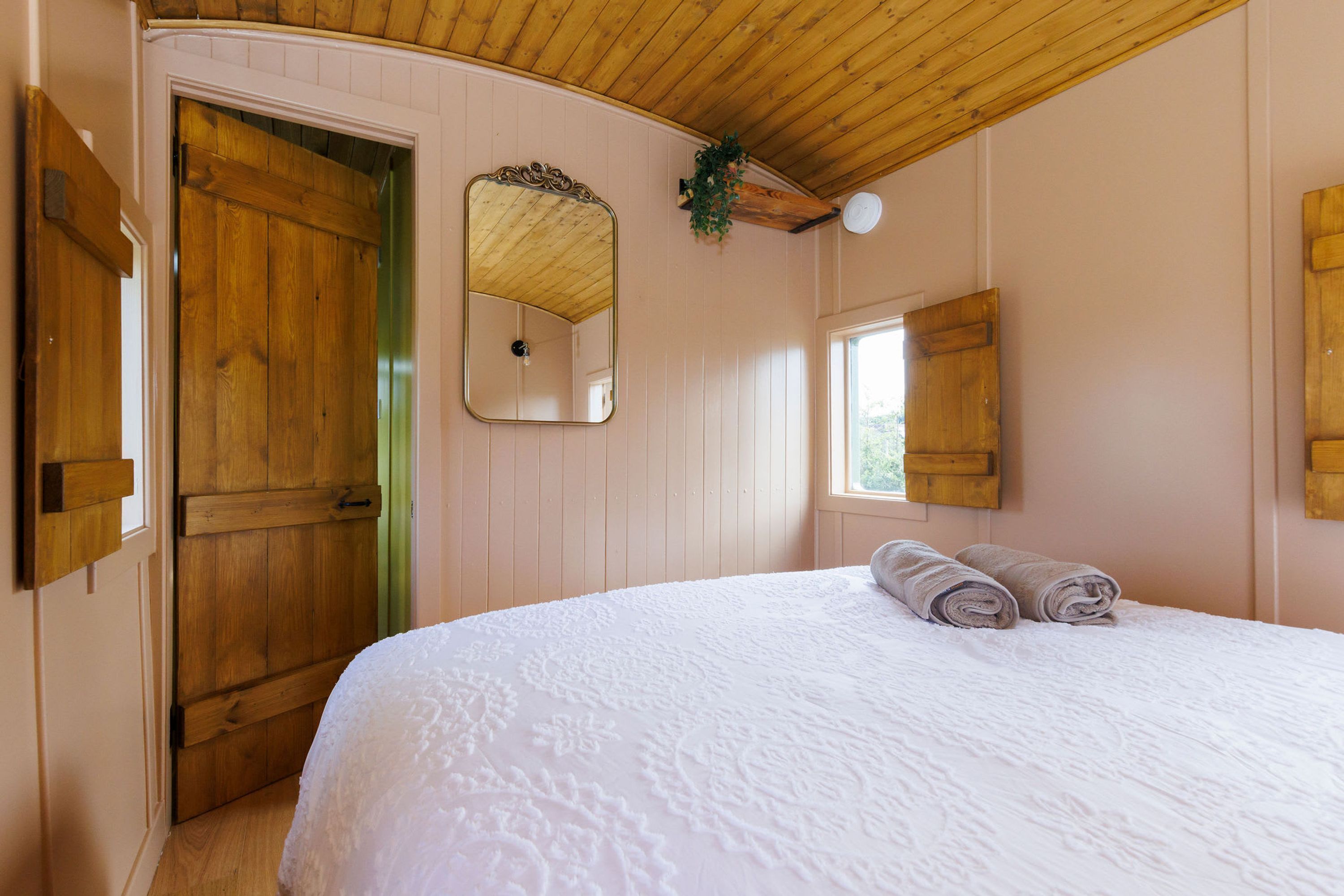 Ein Schlafzimmer mit Holztür, einem Bett mit weißer Bettwäsche, einem Spiegel und einem Fenster mit Vorhängen.