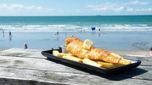 Uma refeição de peixe com batatas fritas sobre uma mesa de madeira, com vista para a praia e o oceano.