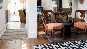 A cozy living room with wooden flooring, a patterned rug, and two wooden chairs with orange cushions. A bookshelf with books and decorative items is visible in the background. - Enchanting 4BD Cottage Lodge Nr Eartham Wood (Petworth)