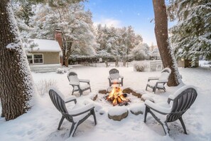 A snow-covered outdoor seating area with a fire pit, surrounded by snow-laden trees and a house in the background.