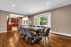 A spacious dining area with a long table and modern chairs, a kitchen with wooden cabinets, and large windows offering a view of the outdoors.
