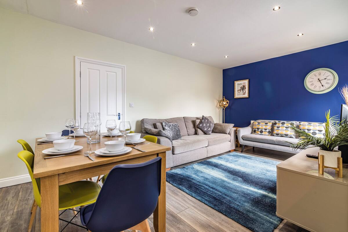 A modern living room with a dining area, featuring a sofa, a coffee table, and a dining table set with plates and glasses.