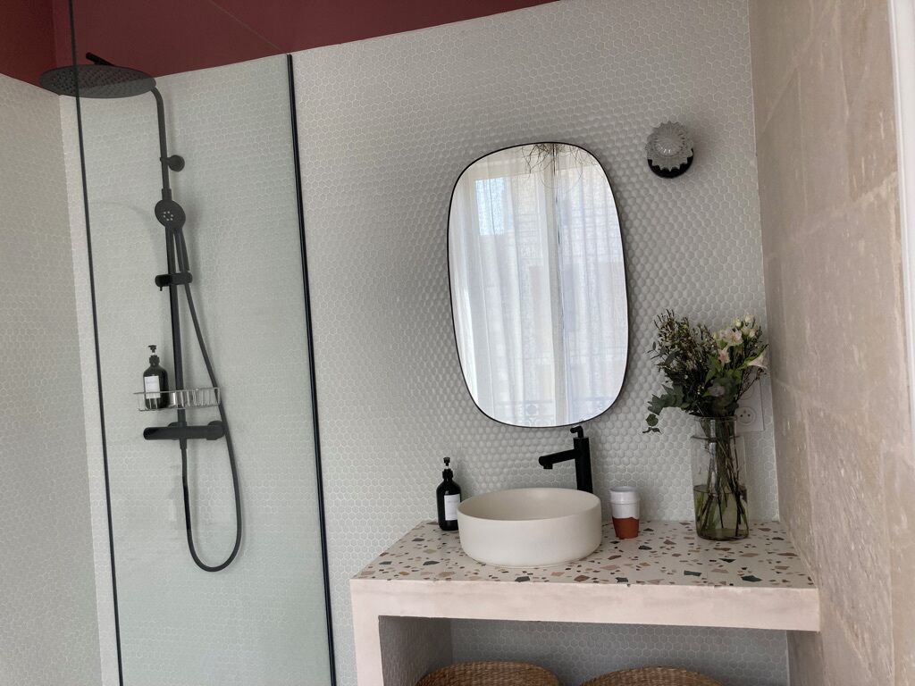 A bathroom with a freestanding shower, a round sink, a mirror, and a vase with flowers.