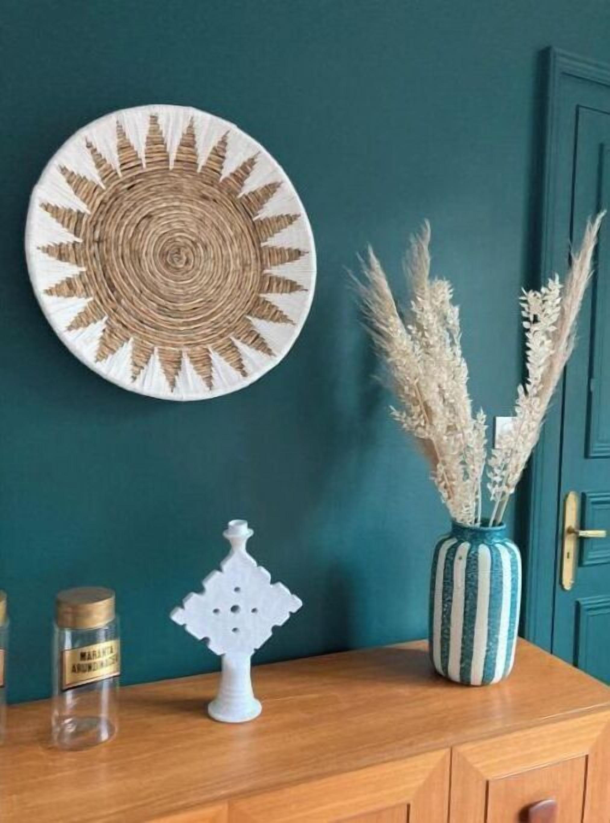 Pared color turquesa con un adorno circular tejido, un jarrón blanco con plantas secas y un pequeño objeto decorativo blanco sobre una superficie de madera.