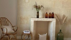 Una habitación con una silla de mimbre, una pequeña mesa redonda con un periódico, un tapete blanco, una cesta de mimbre, un jarrón verde con plumas de pampa y una chimenea con tres botellas marrones encima.