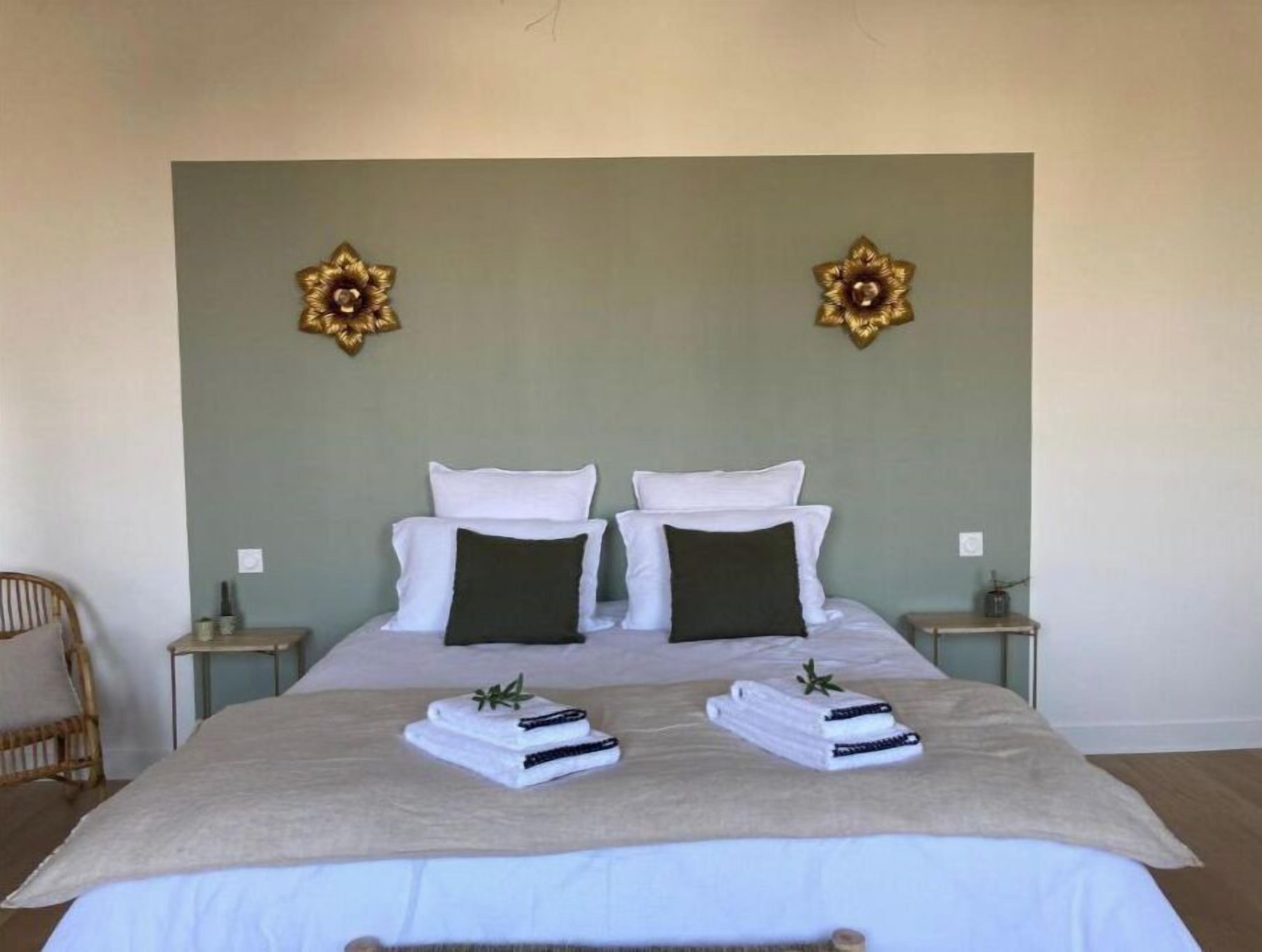 Una cama bien hecha con almohadas blancas y verde oscuro, una manta color beige y dos toallas dobladas con un ramita de verdor encima.
