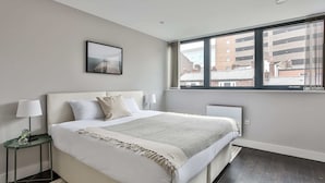 Une chambre moderne avec un grand lit, une table de chevet avec une lampe et une fenêtre donnant sur des immeubles.