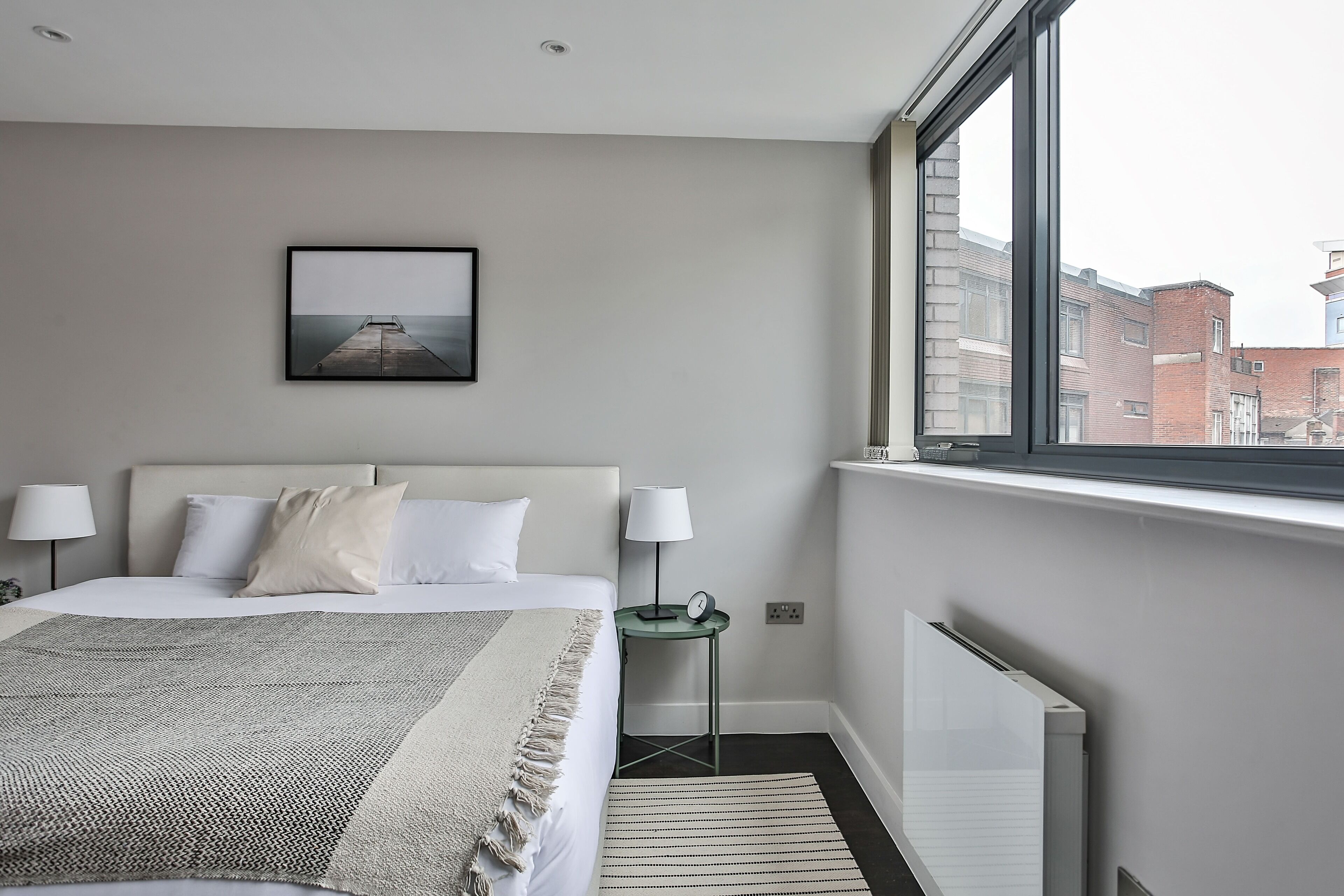 A hotel room with a bed, two bedside lamps, a framed picture on the wall, a small table with a lamp, and a view of buildings through a window.