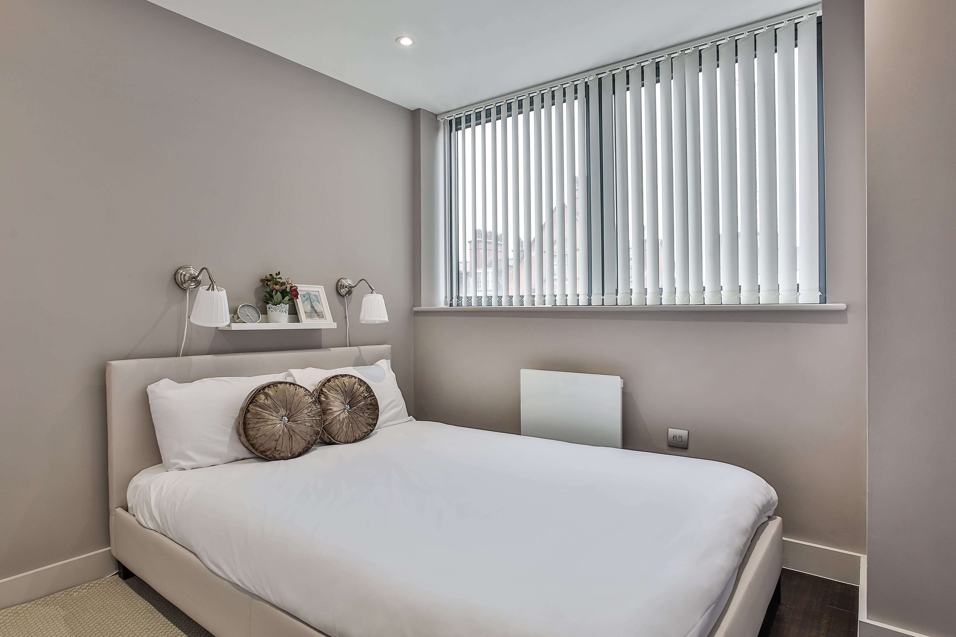 A modern bedroom with a large bed, a bedside table, a wall-mounted shelf with decorative items, and a window with vertical blinds.