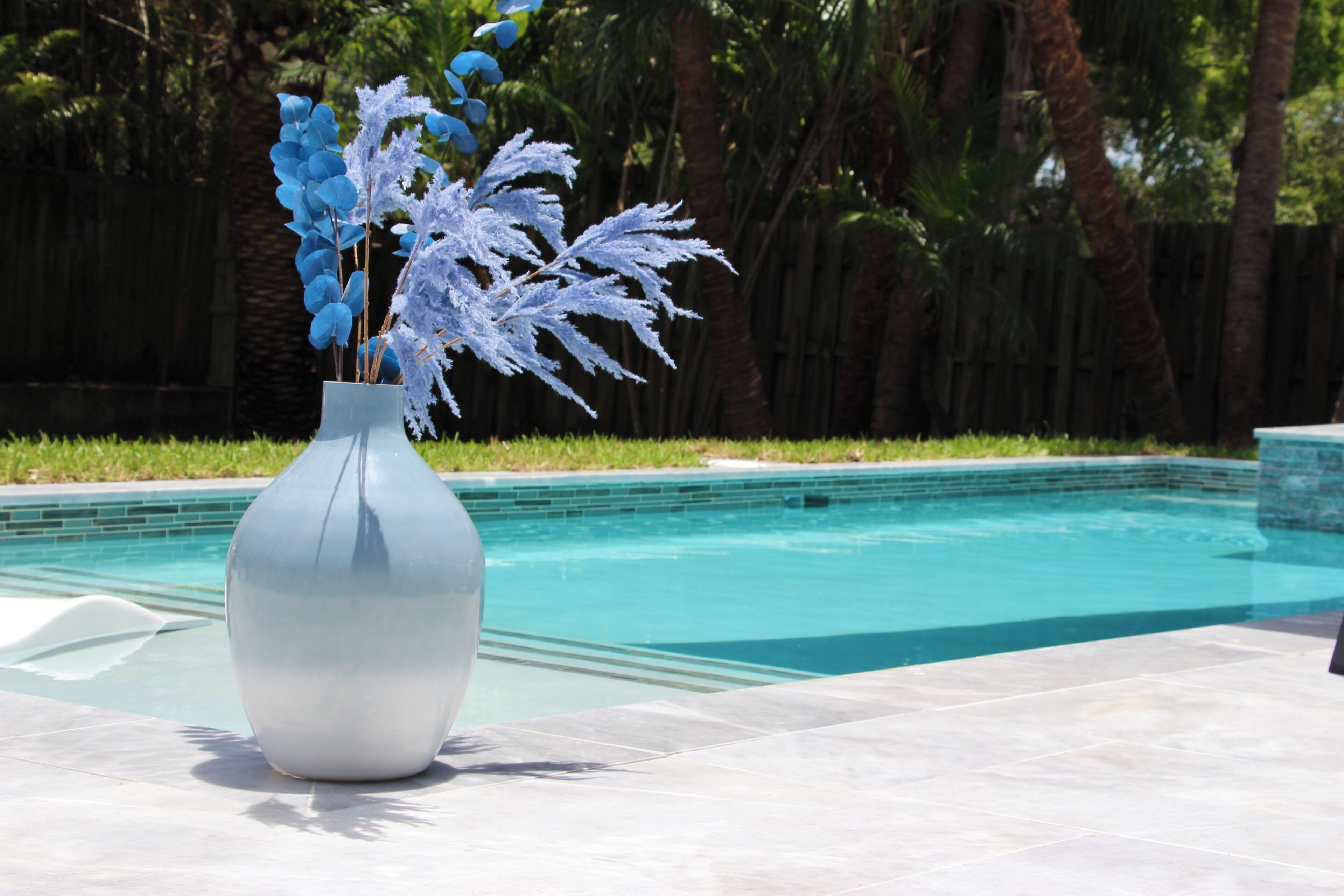 A white vase with blue and white flowers on a tiled patio by a pool.