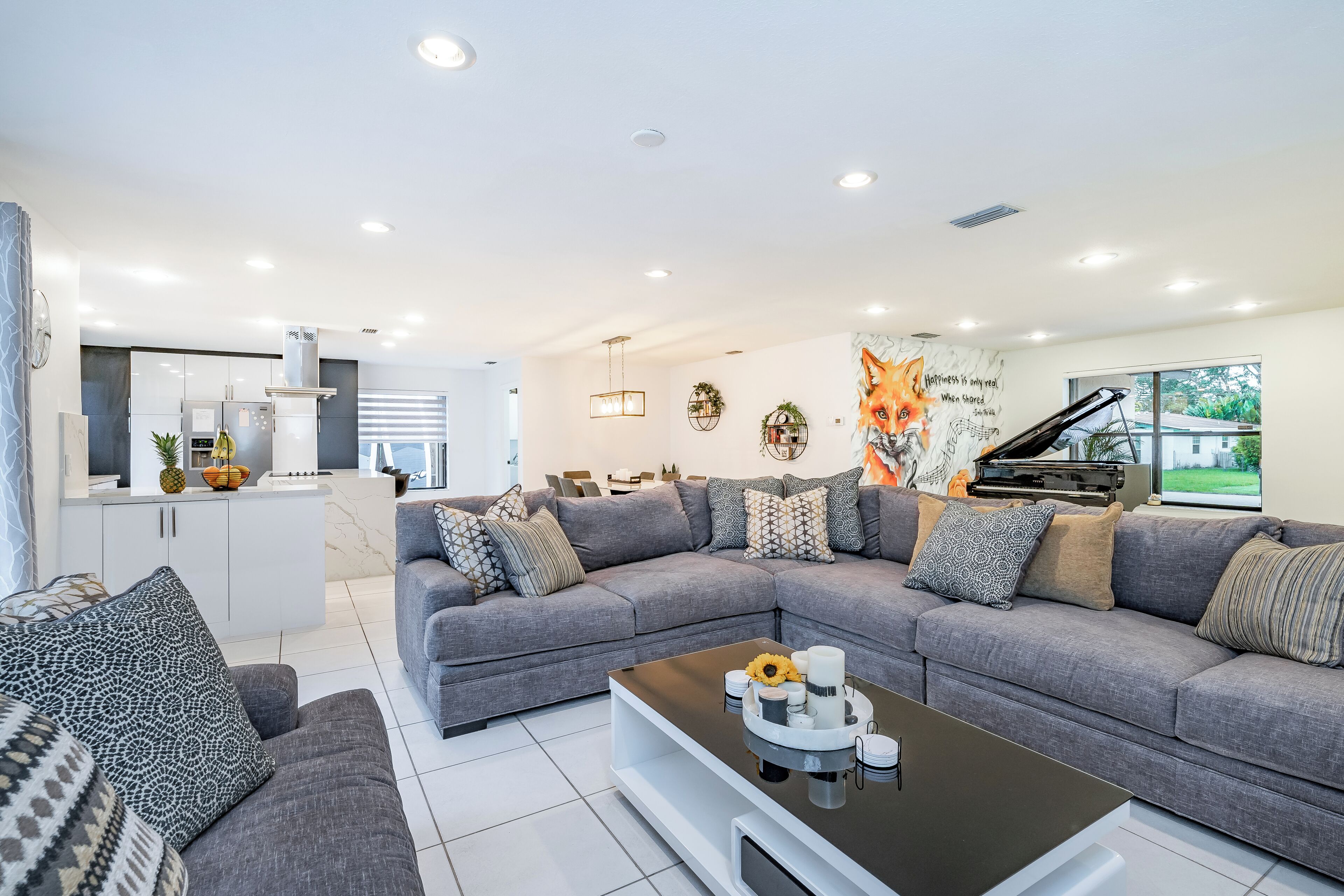 A modern living room with a grey sectional sofa, a coffee table with decorative items, a kitchen area with white cabinets, and a mural of a fox on the wall.