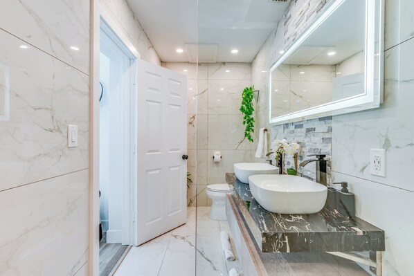 Un baño moderno con baldosas de mármol blanco, un espejo grande, dos lavamanos y un inodoro.