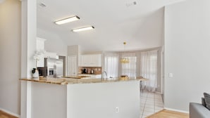 A modern kitchen with a central island, white cabinetry, and a dining area with a view of the living room. - Family Fun Retreat: Pool & Entertainment Haven! 4 Bedroom Home by RedAwning (Frisco)