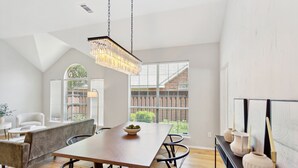 A modern dining area with a wooden table, cushioned bench, and chairs. A chandelier hangs from the ceiling, and there are large windows with blinds. - Family Fun Retreat: Pool & Entertainment Haven! 4 Bedroom Home by RedAwning (Frisco)