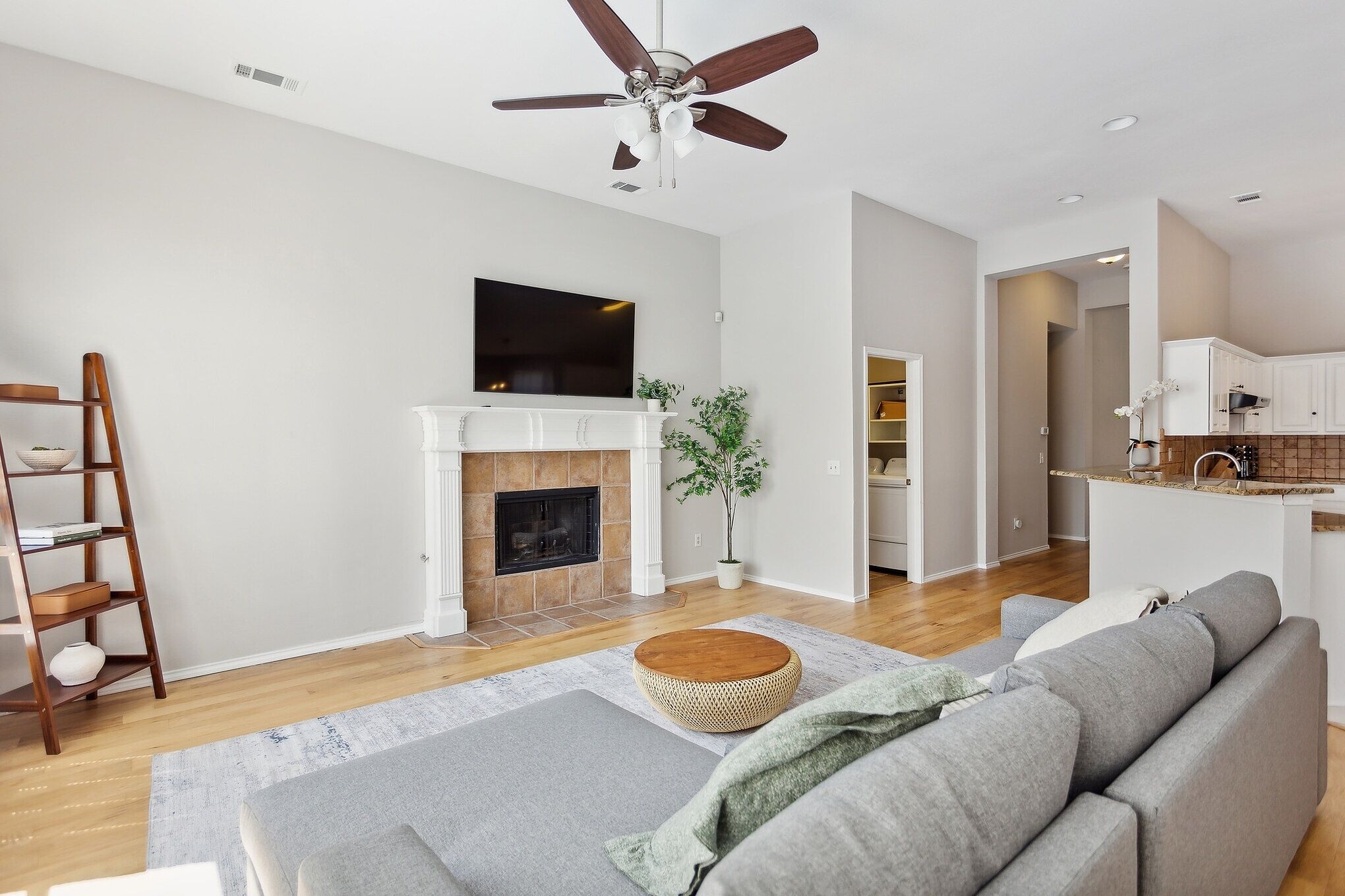 A modern living room with a fireplace, a ceiling fan, a flat-screen TV, and a kitchen area in the background.