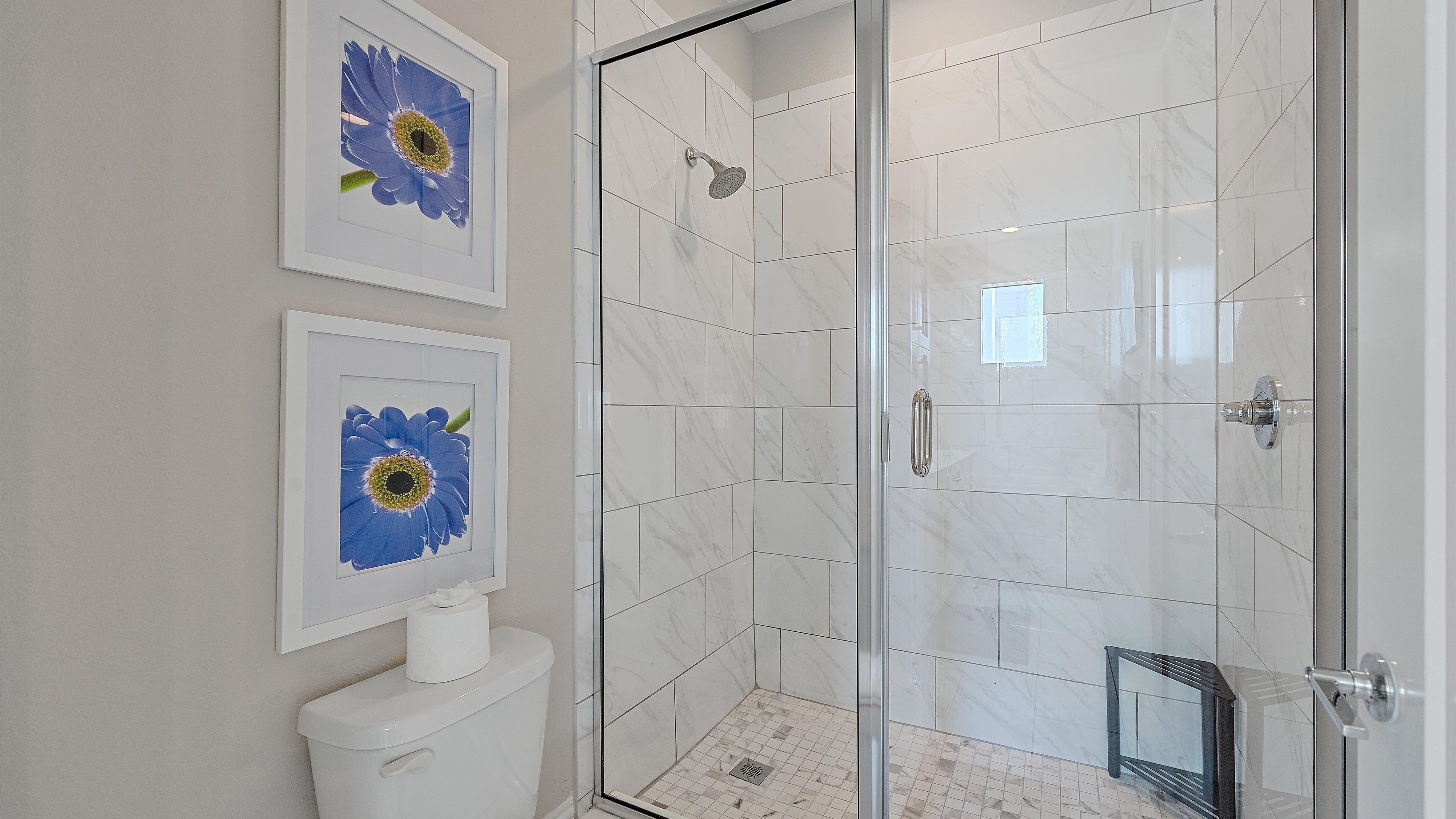 Un bany modern amb una cabina de dutxa de vidre, rajoles de marbre blanc i una obra d'art floral emmarcada a la paret.