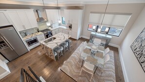 A modern kitchen with white cabinets, a stainless steel refrigerator, a built-in oven, and a large island with a sink. There is a dining area with a glass table and chairs, and a large window providing a view of the outside. - Cityscape Retreat 2 Bedroom Townhouse by RedAwning (Sarasota)