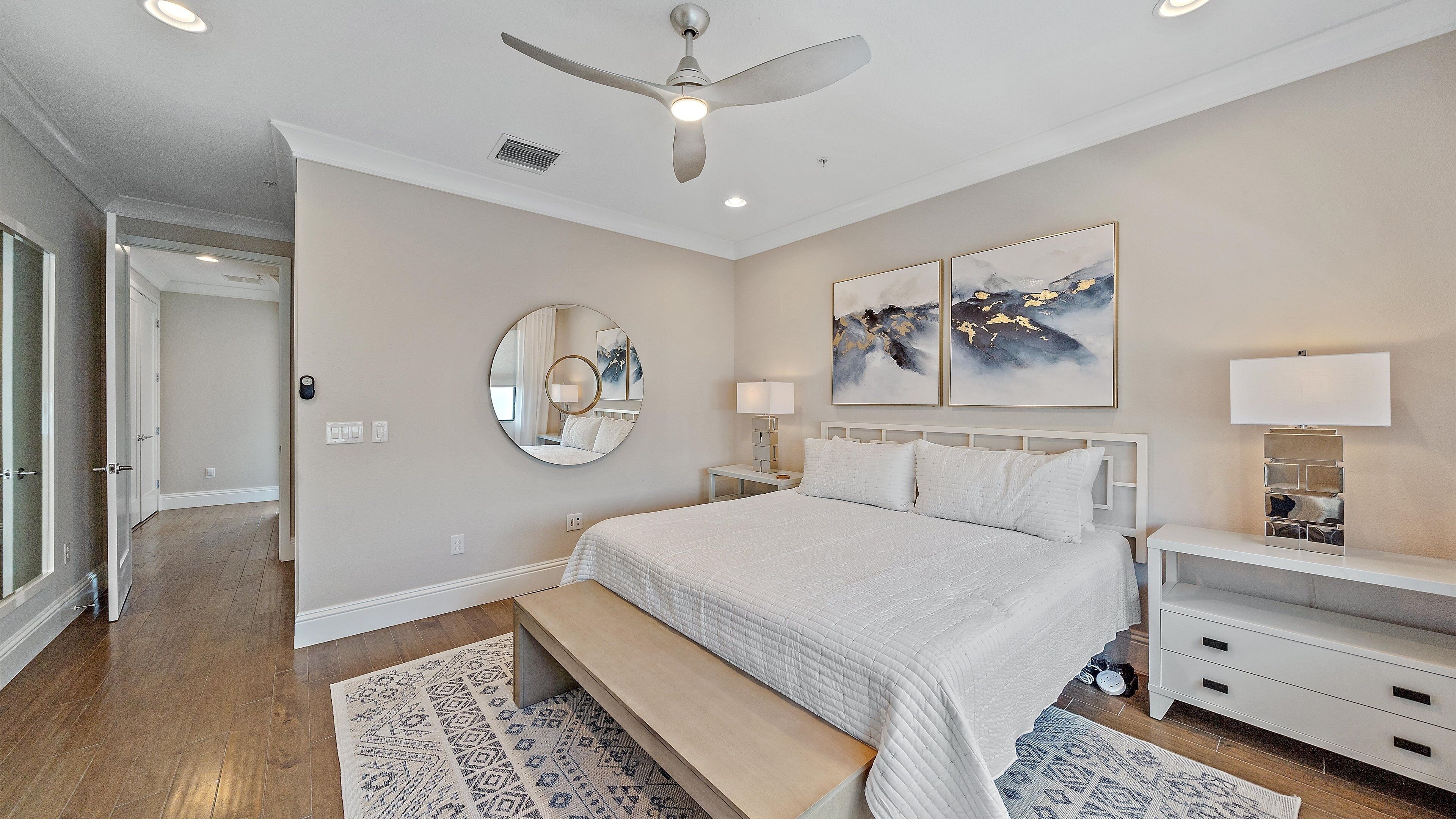 A bedroom with a large bed, a ceiling fan, a bedside table with a lamp, and a wall-mounted TV.
