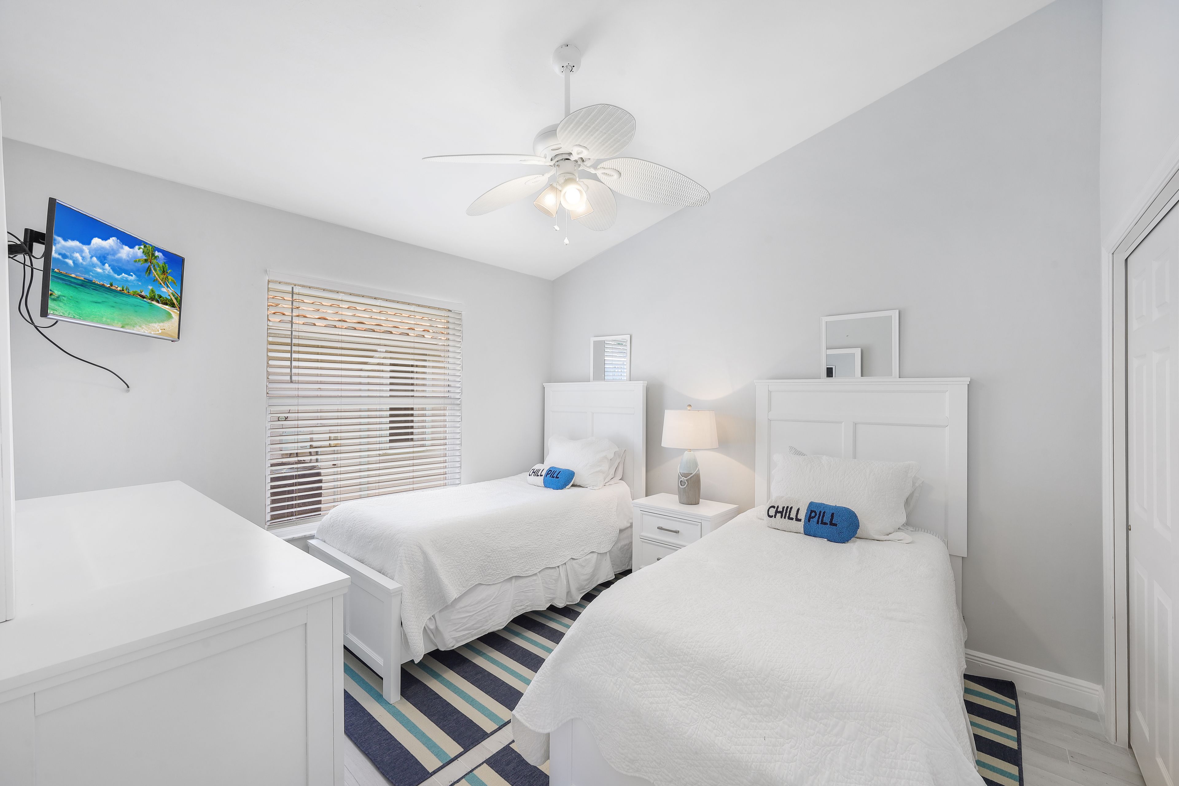 A bedroom with two beds, a ceiling fan, a TV, and a window with blinds.