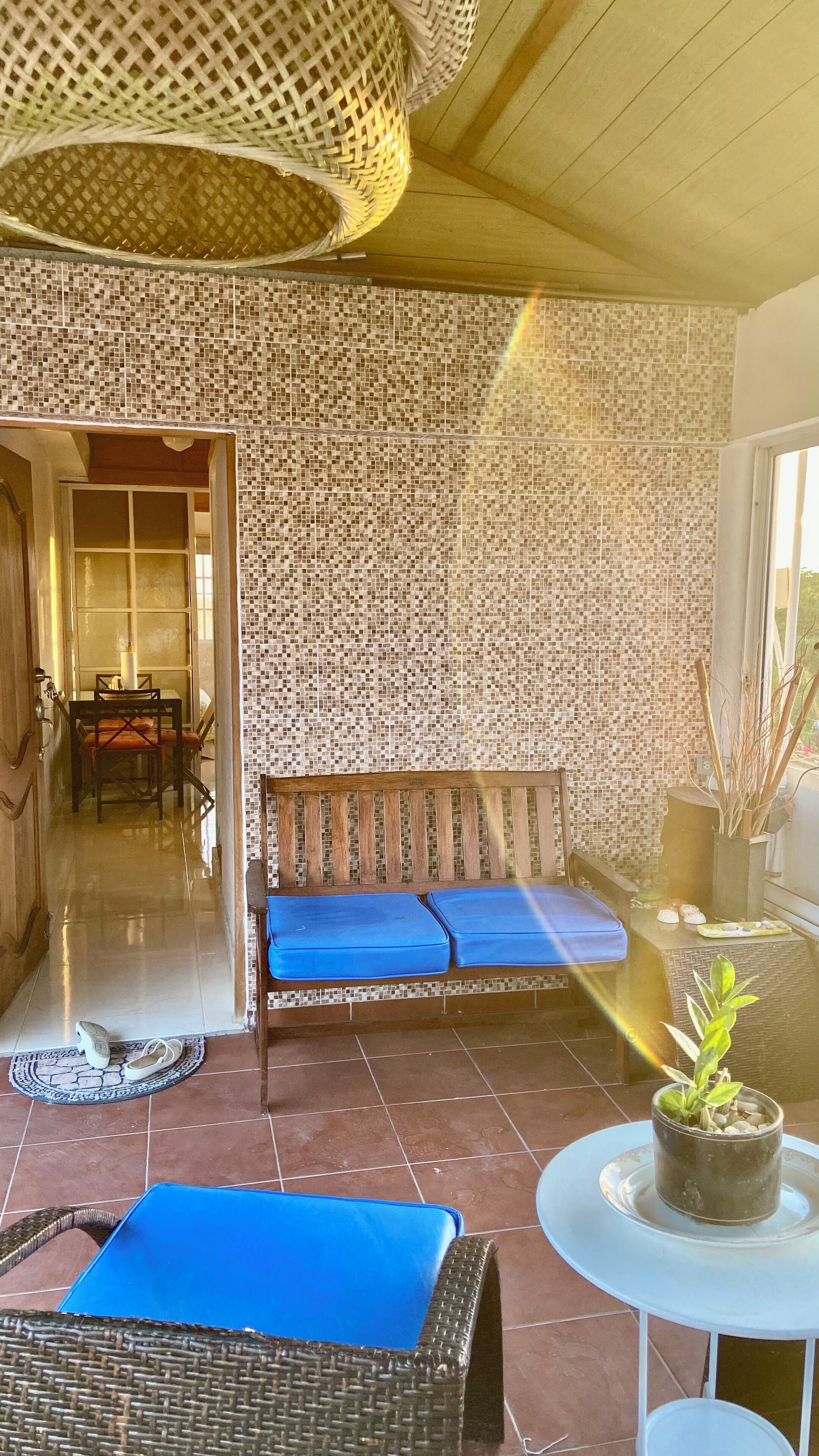 Una habitación con pared de azulejos, un banco de madera con cojines azules, una silla de mimbre, una pequeña mesa con planta en maceta y vistas al exterior a través de una puerta.