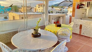 A covered patio with a round table and white wicker chairs, a yellow tablecloth, and a potted plant.