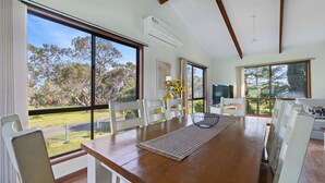A dining area with a wooden table, white chairs, and large windows offering a view of trees and a fence.