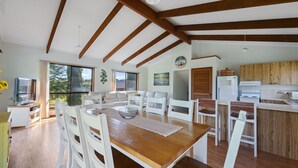 A spacious living room with a wooden dining table, white chairs, and a large window offering a view of greenery. A kitchen with wooden cabinets and a refrigerator is visible in the background.