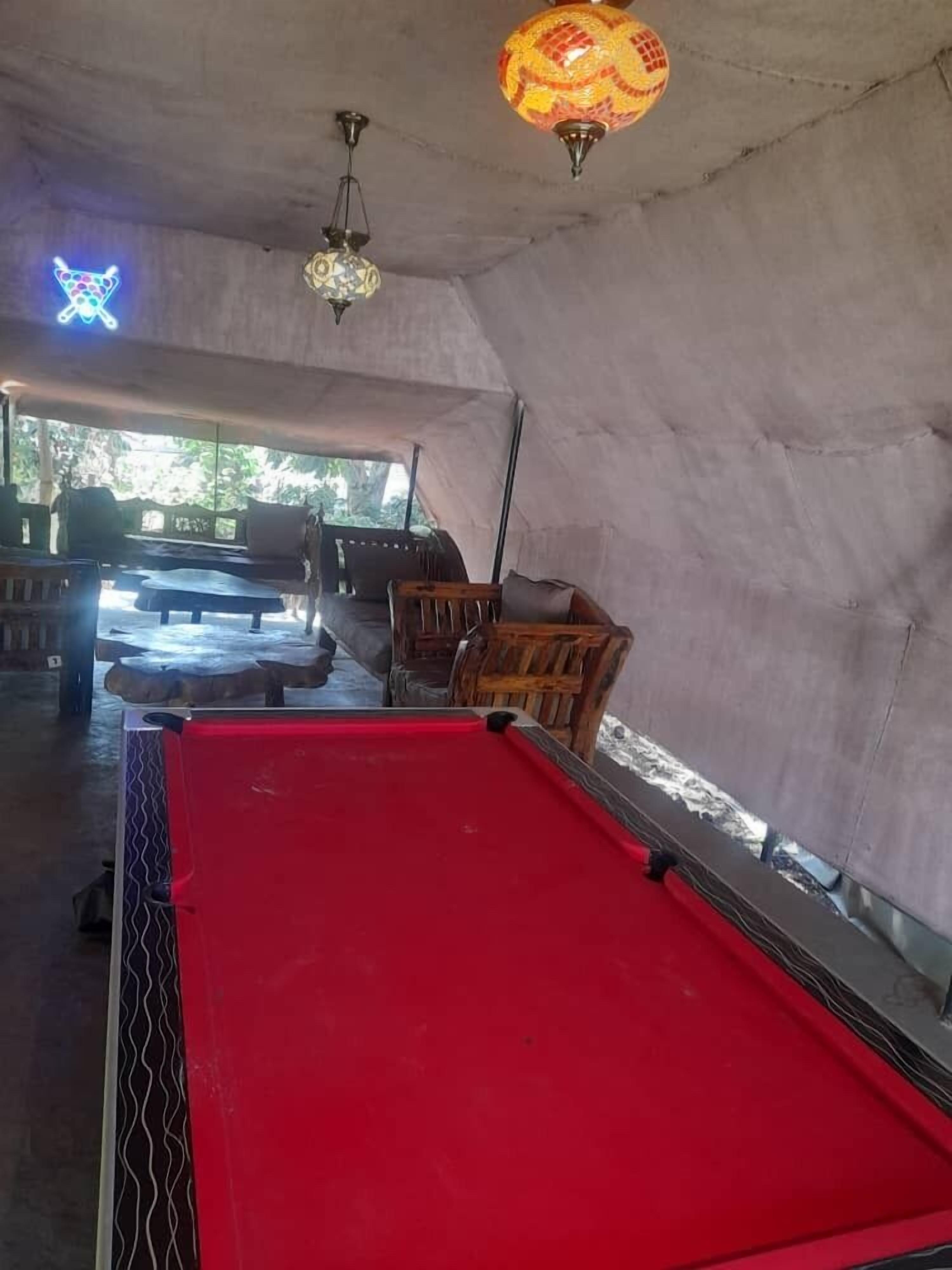 A pool table in a room with a high ceiling and hanging lamps. There are chairs and a sofa in the room.