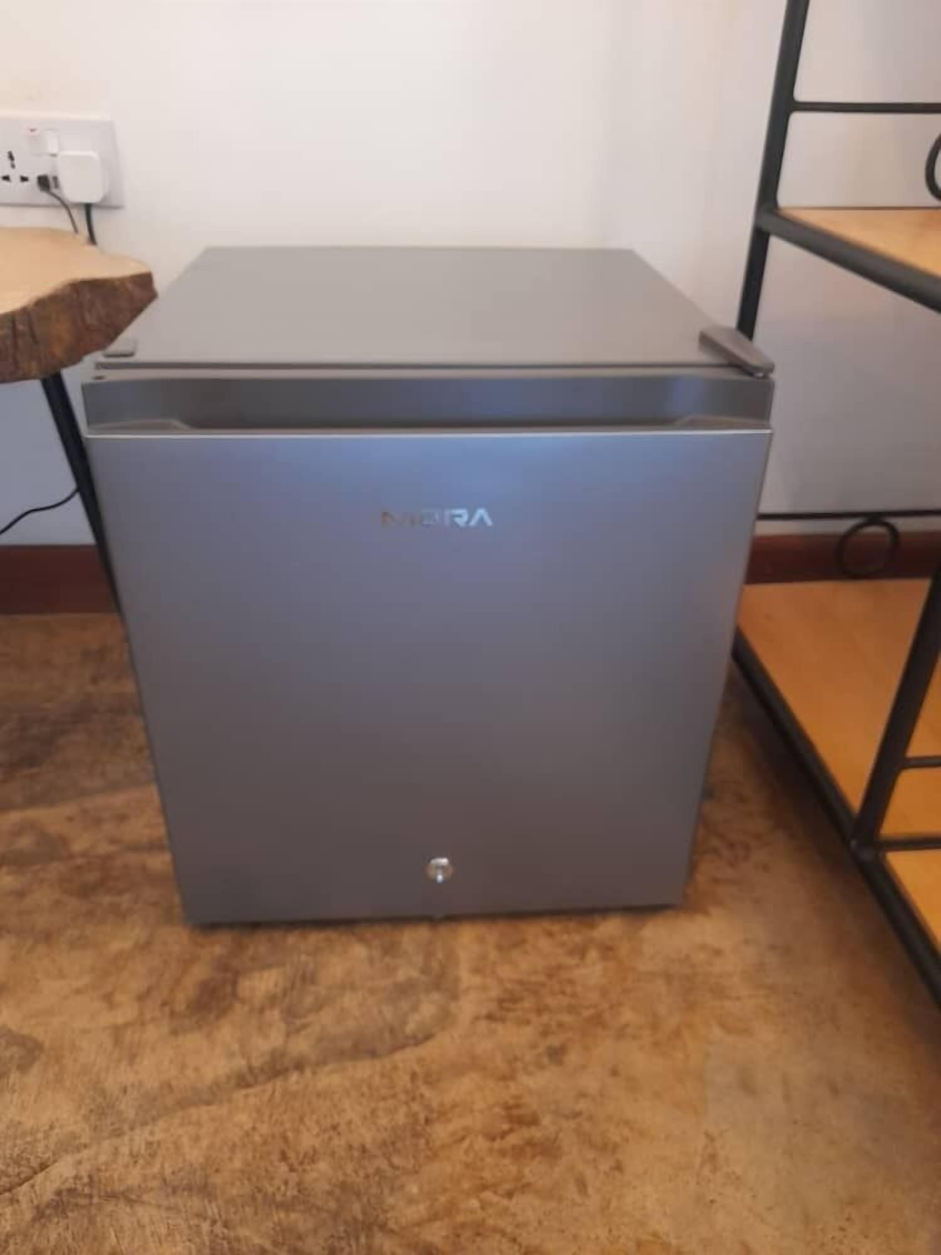 A compact, gray refrigerator with the brand name 'MERA' on the front. It is placed on a wooden floor next to a white wall with a power outlet.