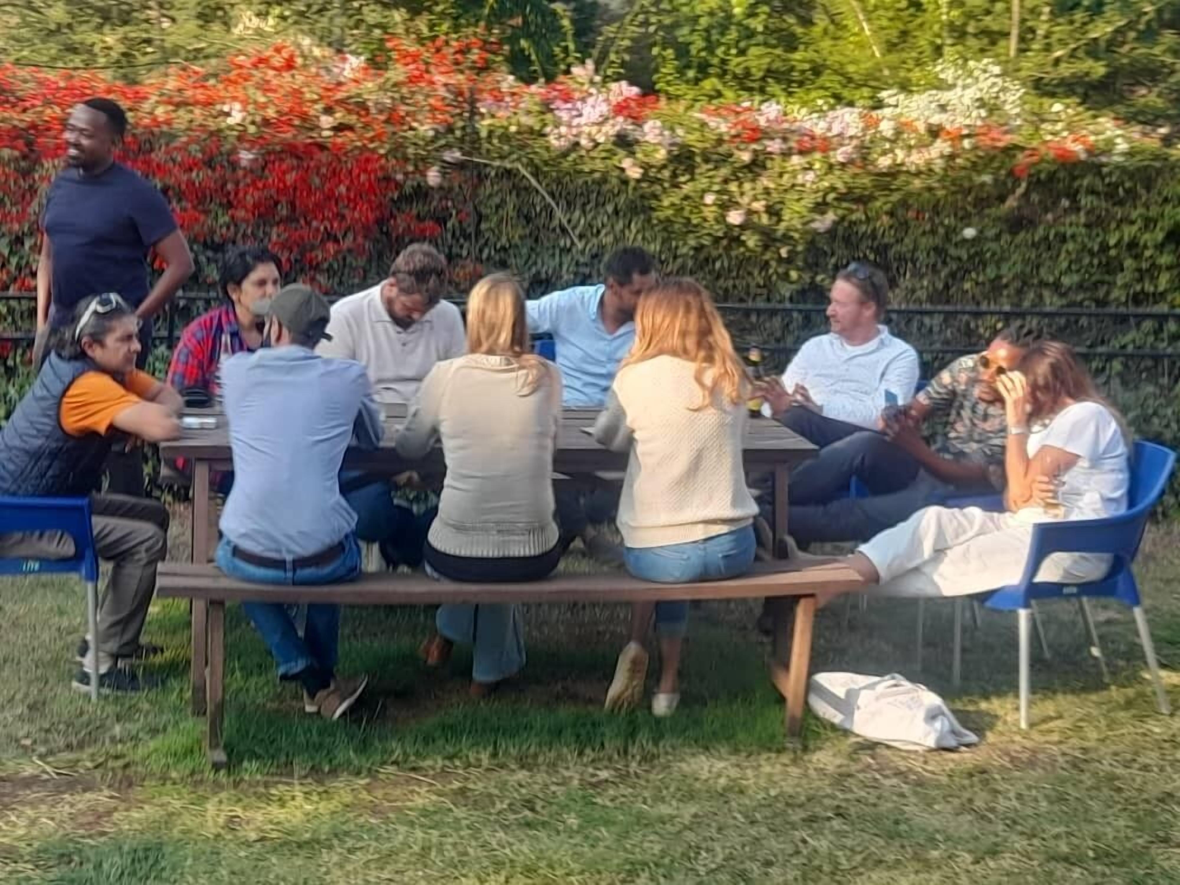 A group of people seated around a wooden table outdoors, with a garden and flowers in the background.