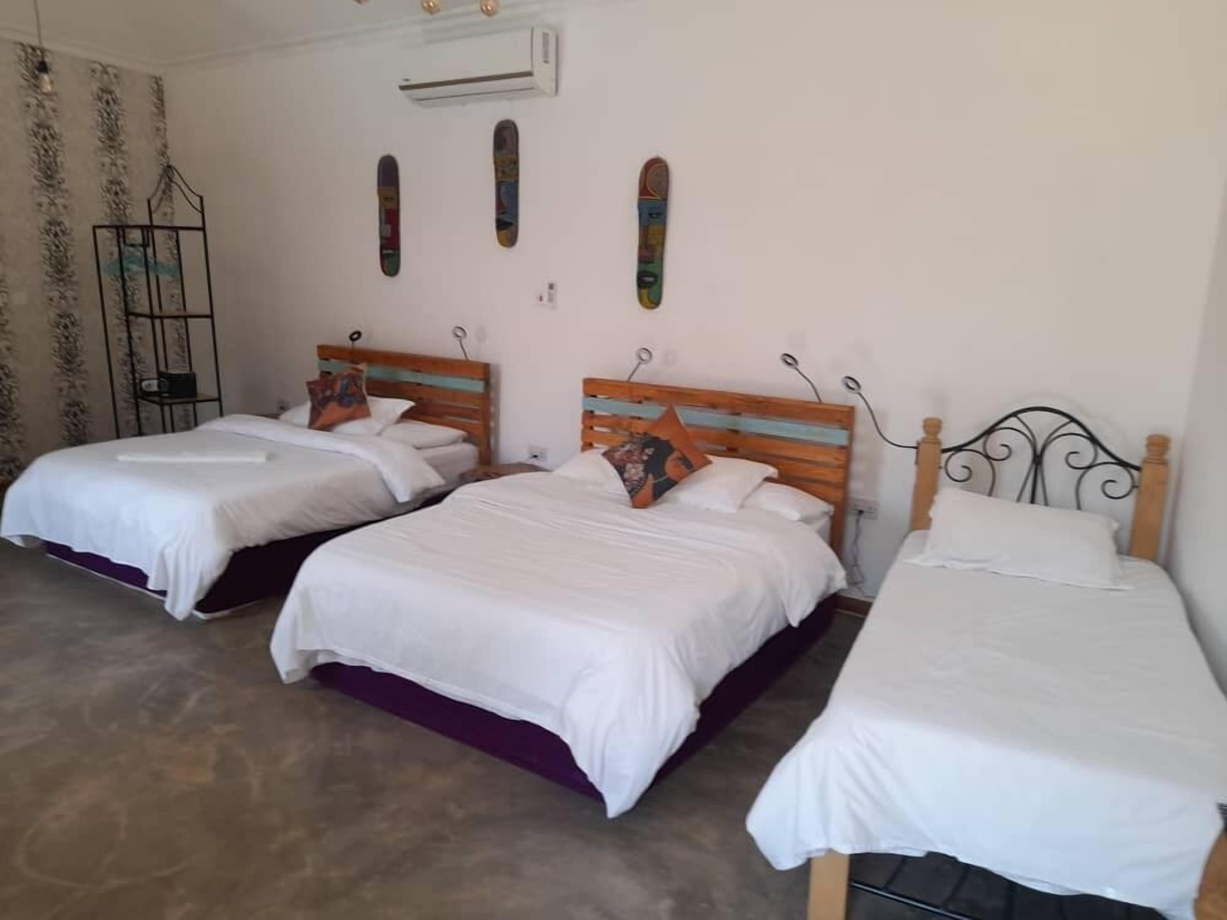 A room with three beds, each with a wooden headboard and white bedding. There is a wall-mounted air conditioner and a small shelf with decorative items.