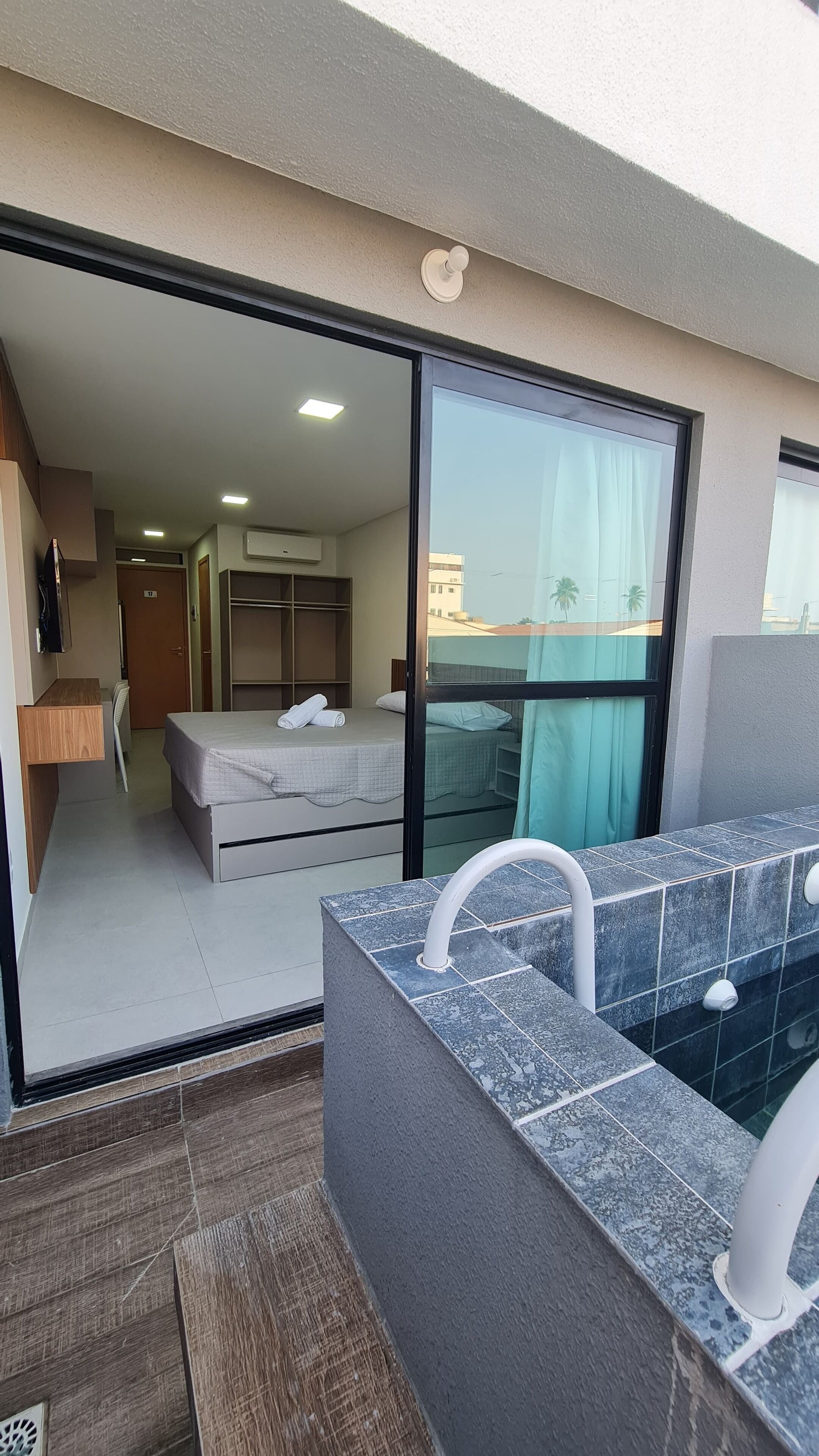 Uma varanda com hidromassagem integrada, uma porta de correr de vidro que dá para um quarto com cama e guarda-roupa, e vista para a paisagem urbana.
