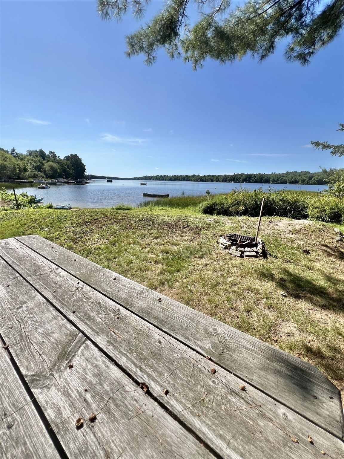 A wooden picnic table in a grassy area with a lake and boats in the background.
