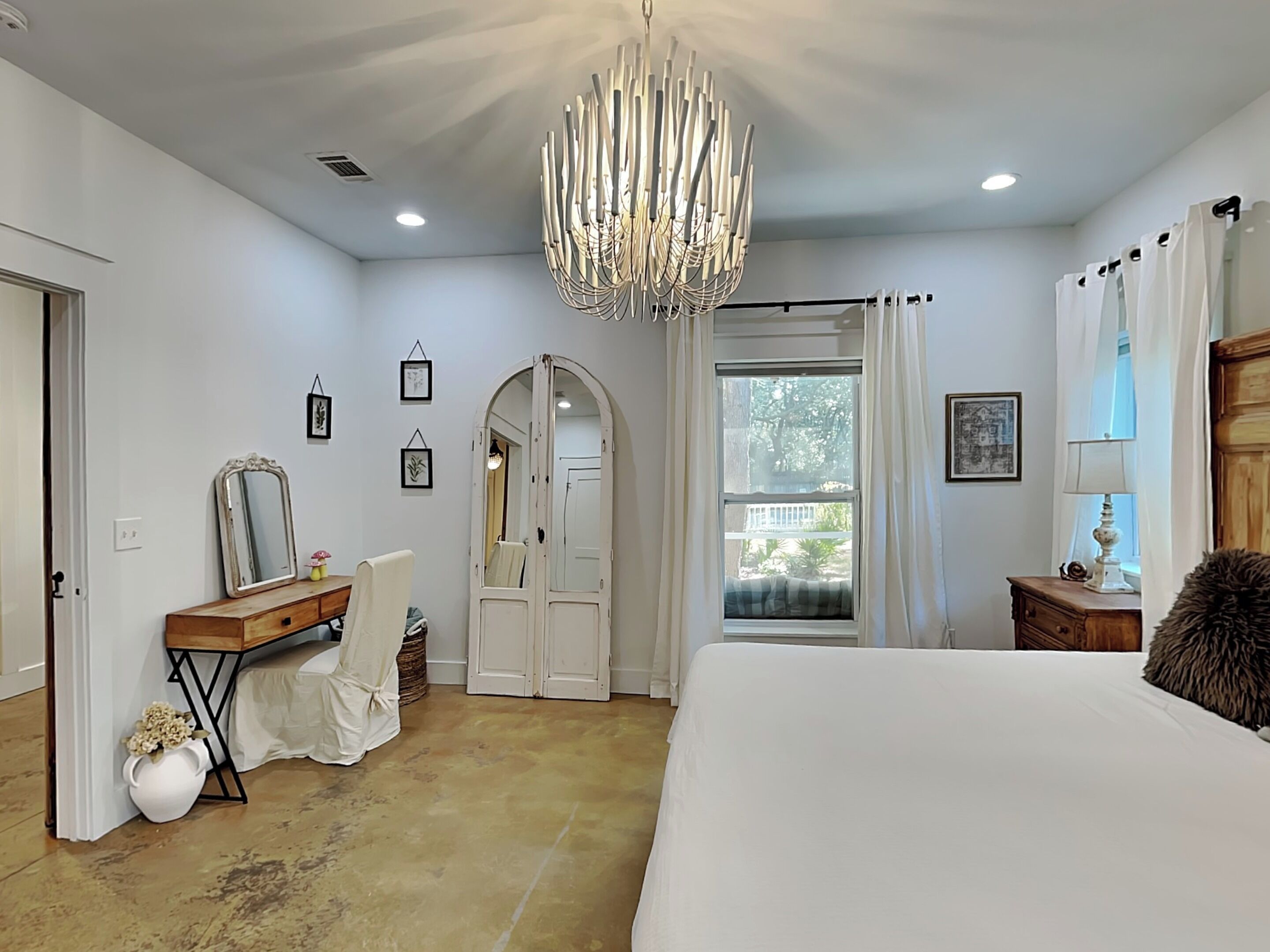A bedroom with a large bed, a vanity with a mirror, a chandelier, and a view of the outdoors through a window.