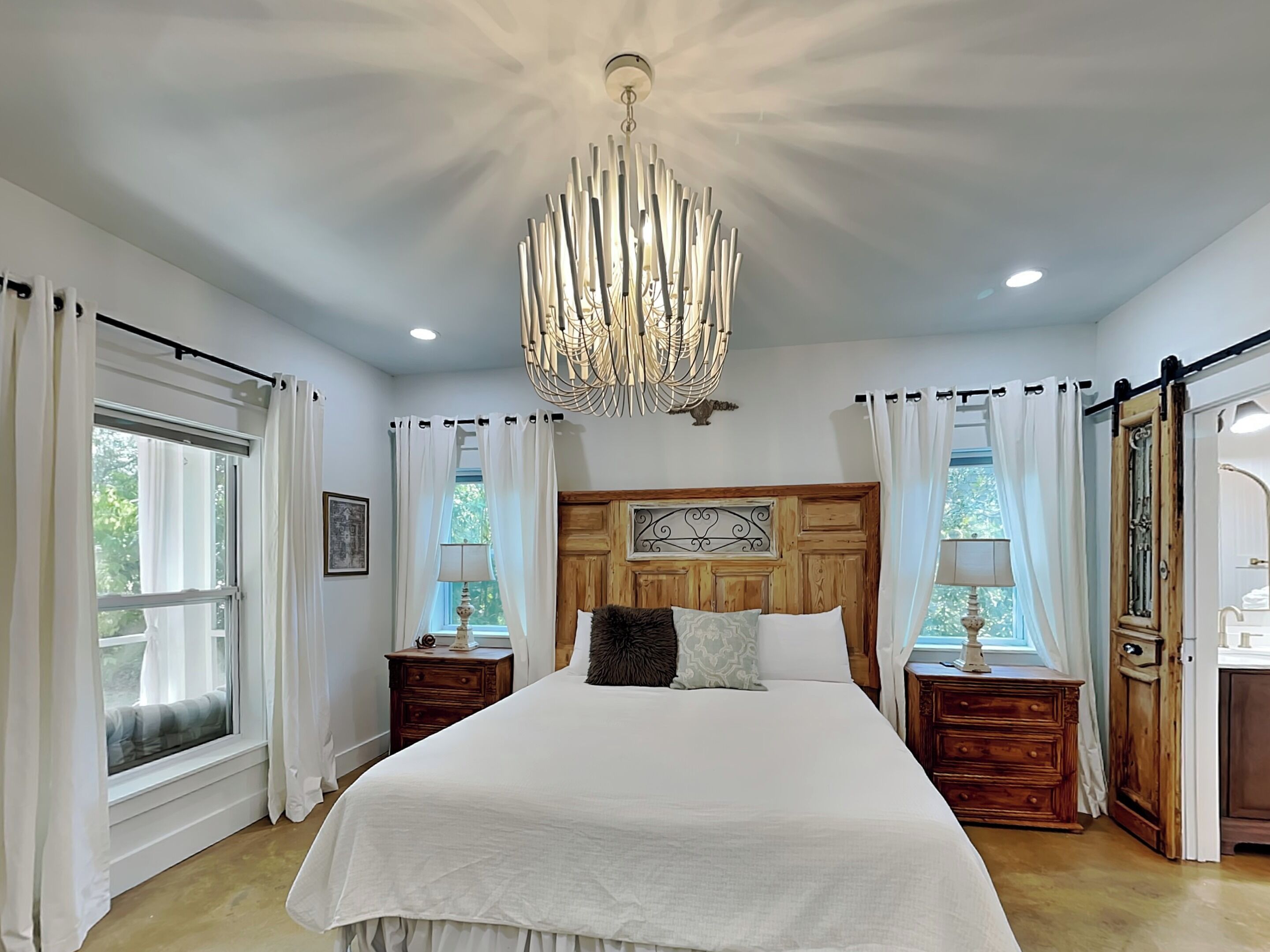 A bedroom with a large bed, wooden headboard, two bedside tables with lamps, a chandelier, and large windows with curtains.