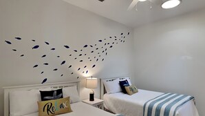 Ein Hotelzimmer mit zwei Betten, jeweils mit einer gestreiften Bettdecke und einem Kissen mit dem Aufdruck 'Relax'. An der Wand ist ein Fisch-Aufkleber angebracht, es gibt einen Deckenventilator und eine Lampe auf einem Nachttisch.