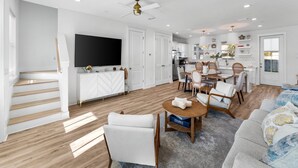 A modern living room with a flat-screen TV, a dining area with a wooden table and chairs, and a kitchen in the background. - Coastal Haven A by Resort Collection (Panama City Beach)