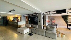 A modern hotel lobby with a reception desk, a seating area with a sofa and a small table, and a wall with a green plant and a sign. - Get One Suites (João Pessoa)