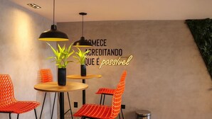 A modern interior with a round wooden table, two orange chairs, black pendant lights, and a wall with a neon sign. - Get One Suites (João Pessoa)