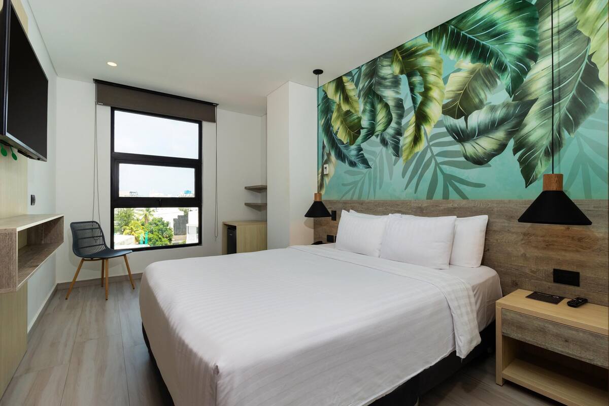 A modern hotel room with a large bed, a desk, a chair, and a large window.