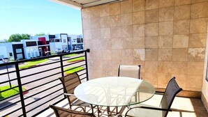 Un balcón con una mesa y sillas de vidrio, vista a un estacionamiento, y un edificio con una puerta roja al fondo.