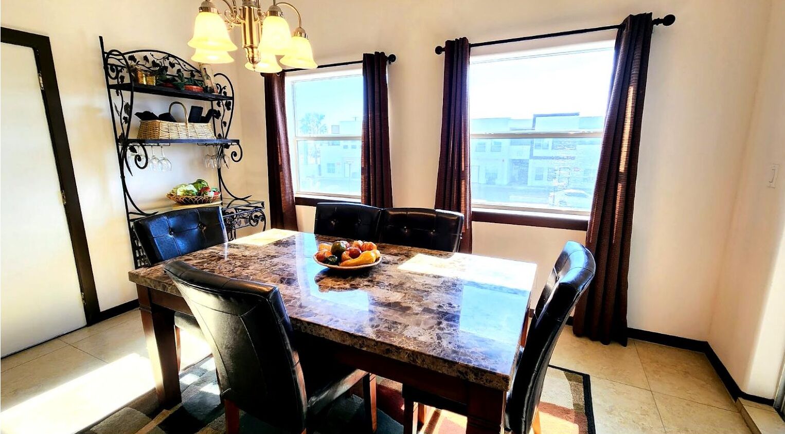 A dining area with a marble table, black chairs, a fruit bowl, and a metal shelf with glassware.