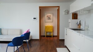 Una cocina moderna con armarios blancos, piso de madera, zona de comedor con sofá blanco y sillas azules, y una pequeña mesa amarilla con objetos decorativos.